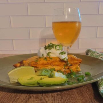 plated portion of finished chicken enchilada casserole topped with sour cream, green onion, and cilantro. lime wedge and chopped fresh avocado on the side, golden pale lager in glass behind