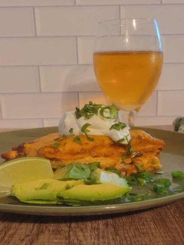 plated portion of finished chicken enchilada casserole topped with sour cream, green onion, and cilantro. lime wedge and chopped fresh avocado on the side, golden pale lager in glass behind