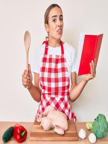cook in apron in front of a raw chicken holding a spoon in one hand and a recipe book in the other, looking confused and disgusted