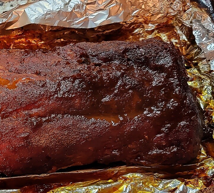 close up of finished rack of barbecue baby back ribs, showing dark bronzed bark and juices, in open foil