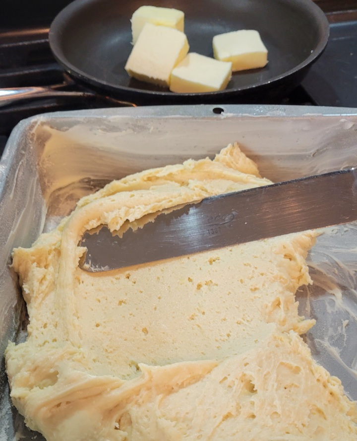 foreground; batter being smoothed into well buttered pan with offset spatula, background; skillet on stove containing butter chunks