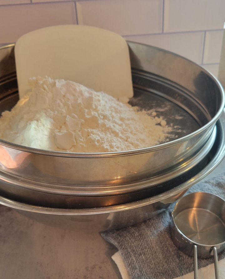 drum sieve over mixing bowl with plastic dough scraper leaning against side, dry ingredients waiting to be sifted