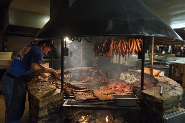 a man seen cooking over a very large round open barbecue pit with multiple items seen cooking.
